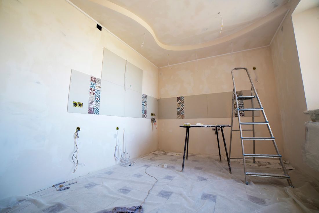 An empty room mid-renovation with bare plaster walls, a stepladder, and protective sheeting on the floor