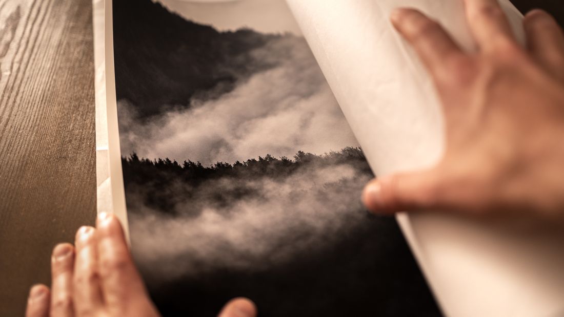Close angled view of hands peeling back a protective paper sleeve from a black-and-white printed landscape photograph on a wood table.