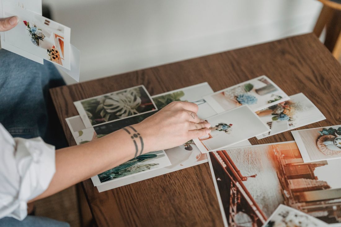 Overhead view of a hand sorting a spread of overlapping printed photographs across a wooden table.