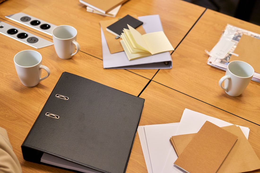 A shared meeting table with a document binder, notebooks, and several coffee mugs set out for a multi-person review