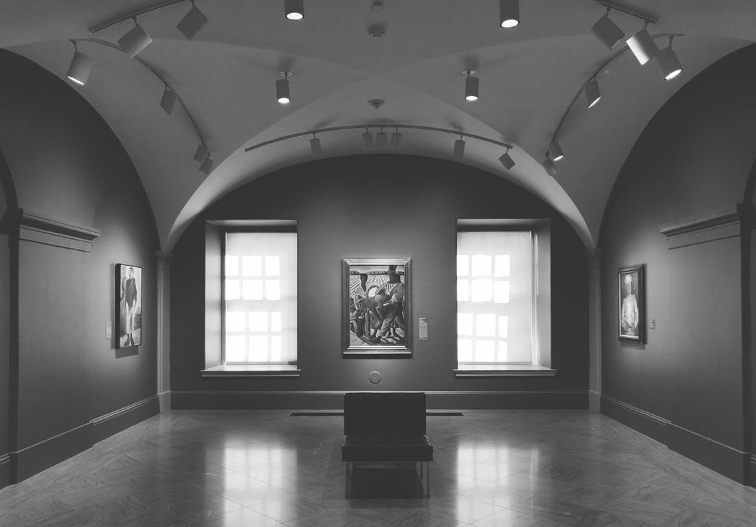 An empty gallery room with vaulted ceilings, framed artwork on the walls, and a single bench