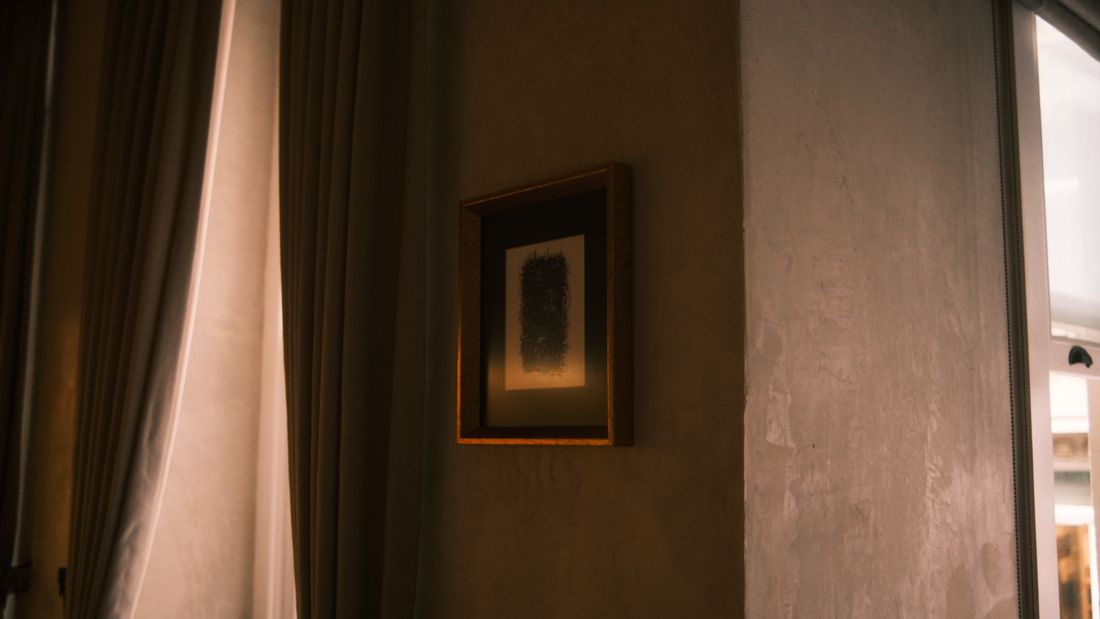 A small framed piece of art hanging on a wall between heavy curtains near a window, warm ambient light
