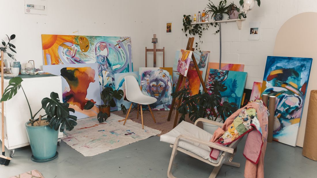 Several large canvases from the same body of work leaning together against a studio wall, plants and a chair in the room