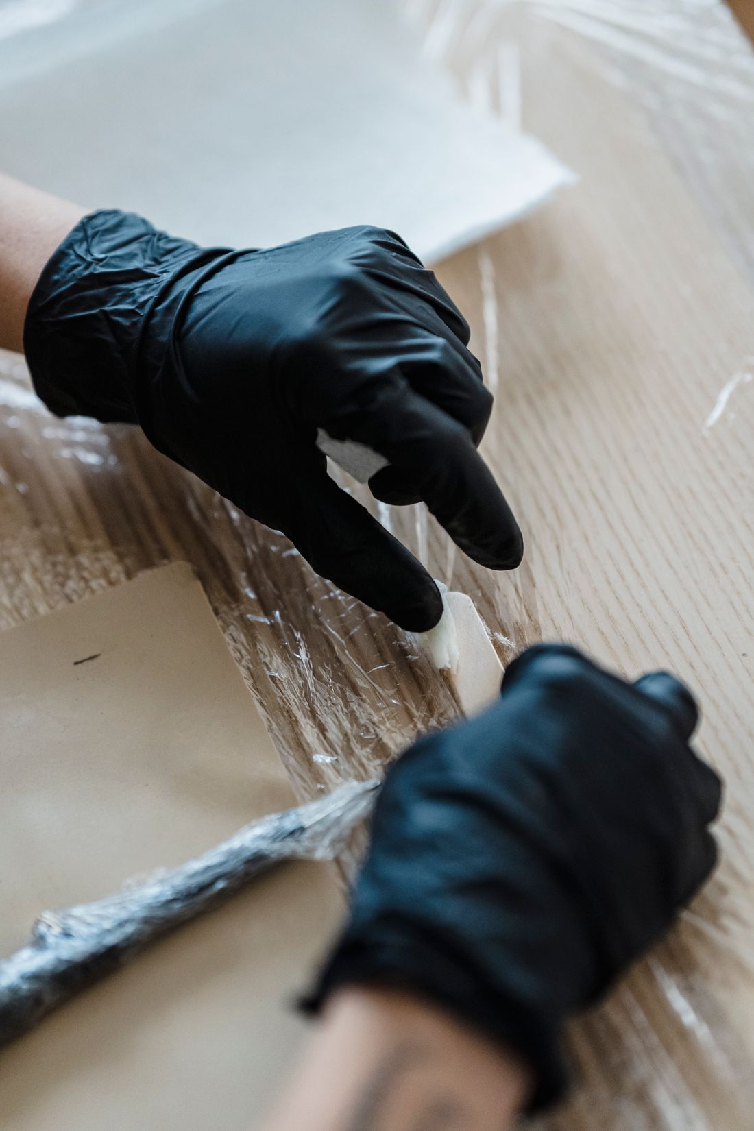 A pair of gloved hands carefully adjusting clear plastic wrap around the taped edge of a wrapped panel on a wooden table