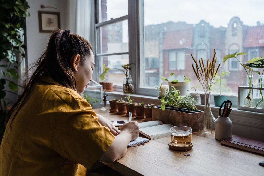 A person seated at a desk beside a window, writing in a notebook, city rooftops visible through the glass