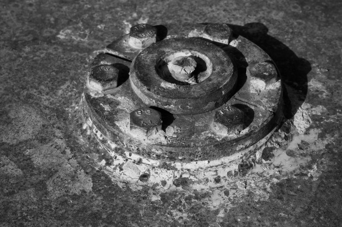 Close-up black-and-white view of a large weathered bolted metal flange fixed into a stone surface, showing the physical construction of the mounting.