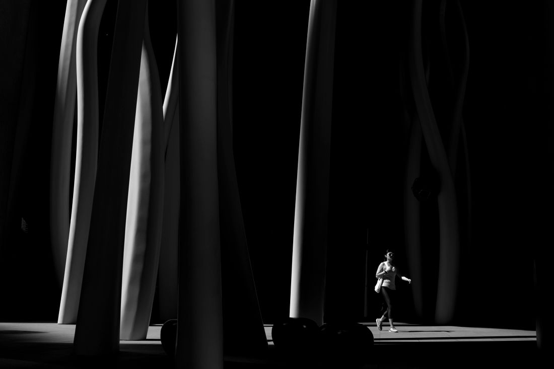 A single figure, face not visible, walks through a row of tall curved sculptural forms lit dramatically in black and white.
