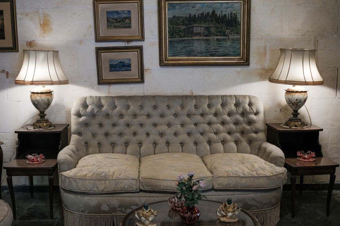 A cream tufted sofa flanked by two lit table lamps beneath a cluster of framed landscape paintings hung close together on a stained plaster wall