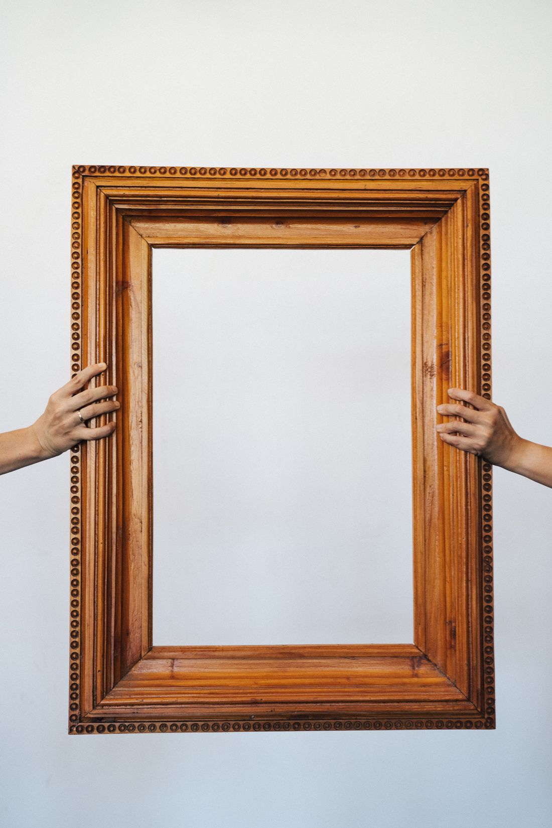 Two hands steady the sides of a large ornate wooden picture frame held up against a plain white wall