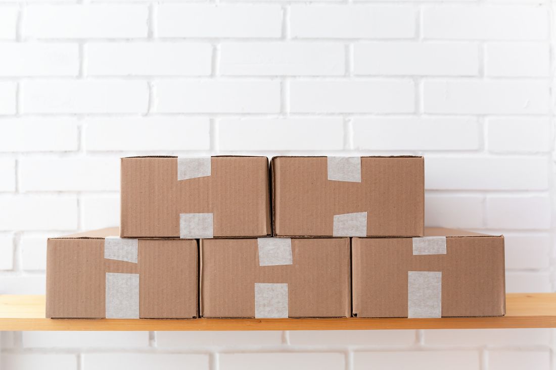 A stack of unlabeled cardboard boxes with plain tape on a wooden shelf against a white brick wall
