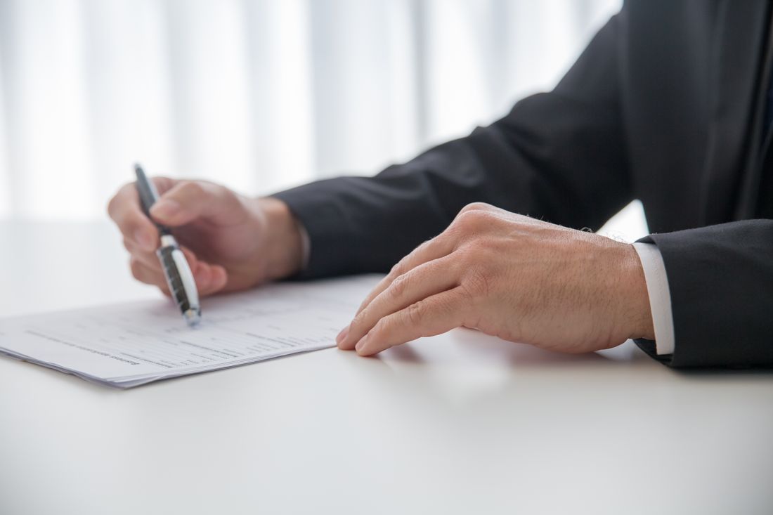 A close view of two hands at a desk, one holding a pen and writing on a printed page angled so the text is not readable