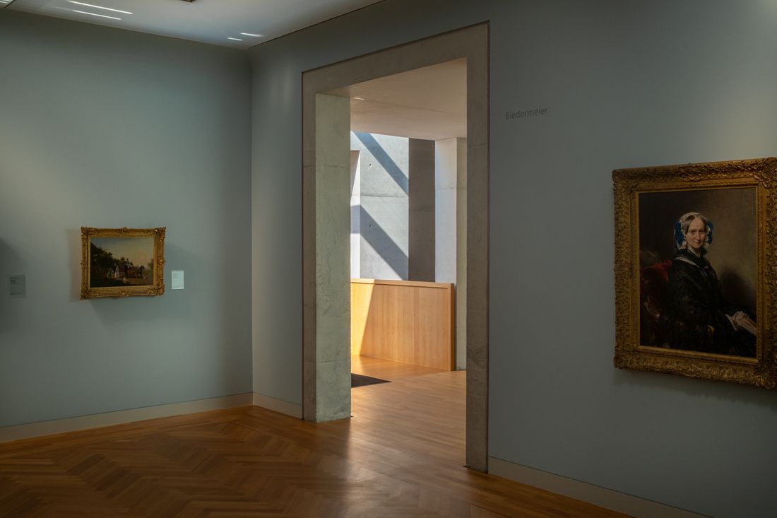 A gallery room seen through an open doorway into an adjacent space, with framed paintings visible on both walls