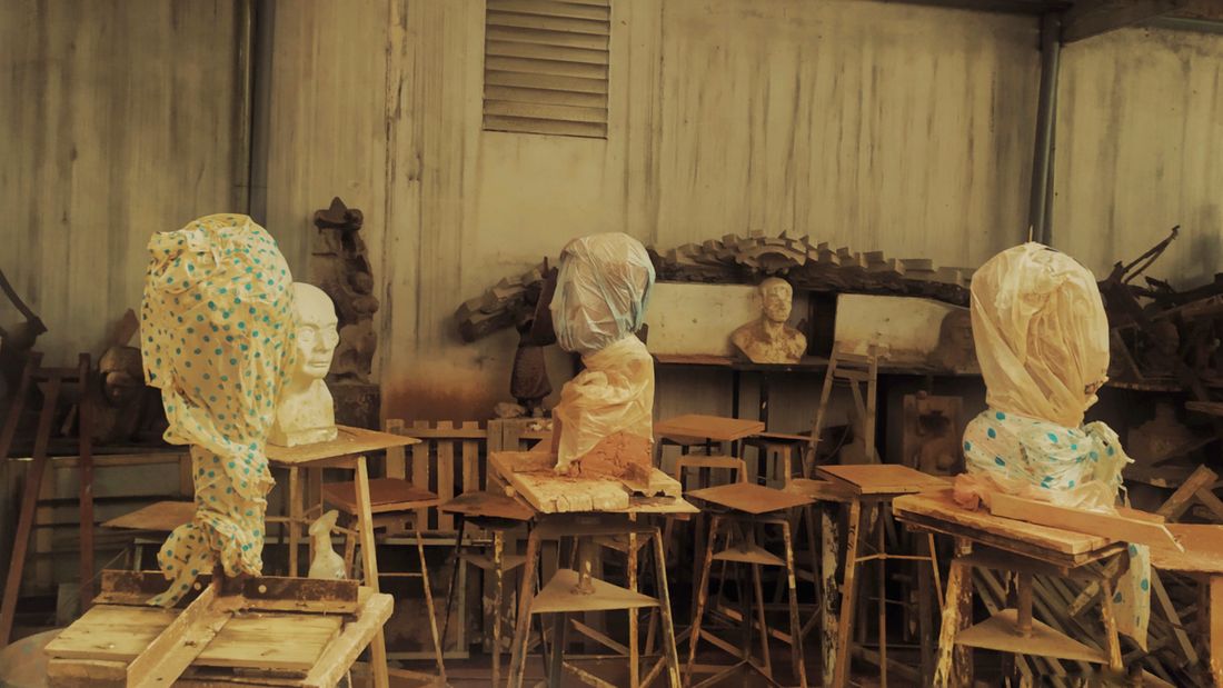 Three plaster sculpture busts on wooden stands in a workshop, each head wrapped and covered in cloth or plastic sheeting for protection.