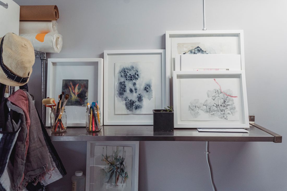 Several small framed paintings and works on paper resting on a wall-mounted shelf next to jars of paintbrushes, with wire shelving alongside.