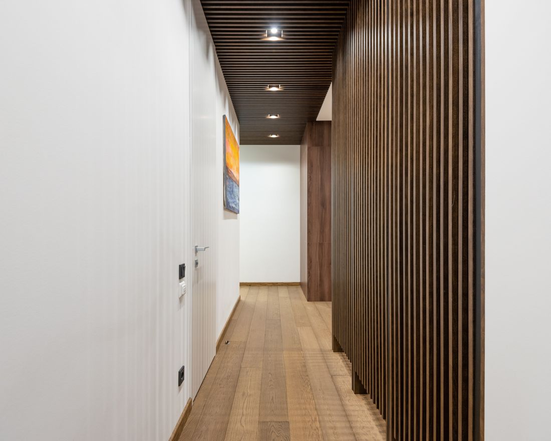 A narrow, plainly finished hallway with white walls, wood paneling, and a closed door at the far end, with one small painting hanging along the wall.