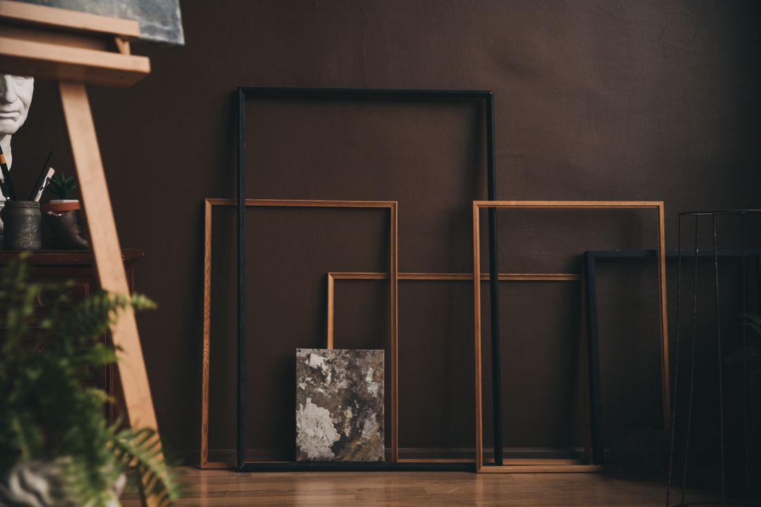 Several empty picture frames of different sizes and one small textured abstract canvas leaning against a dark studio wall, with a wooden easel nearby and no people in the frame.