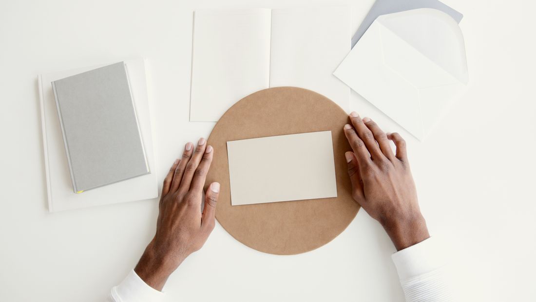 Two hands arranging a blank card and envelope on a round kraft-paper board on a plain white table, seen from above.