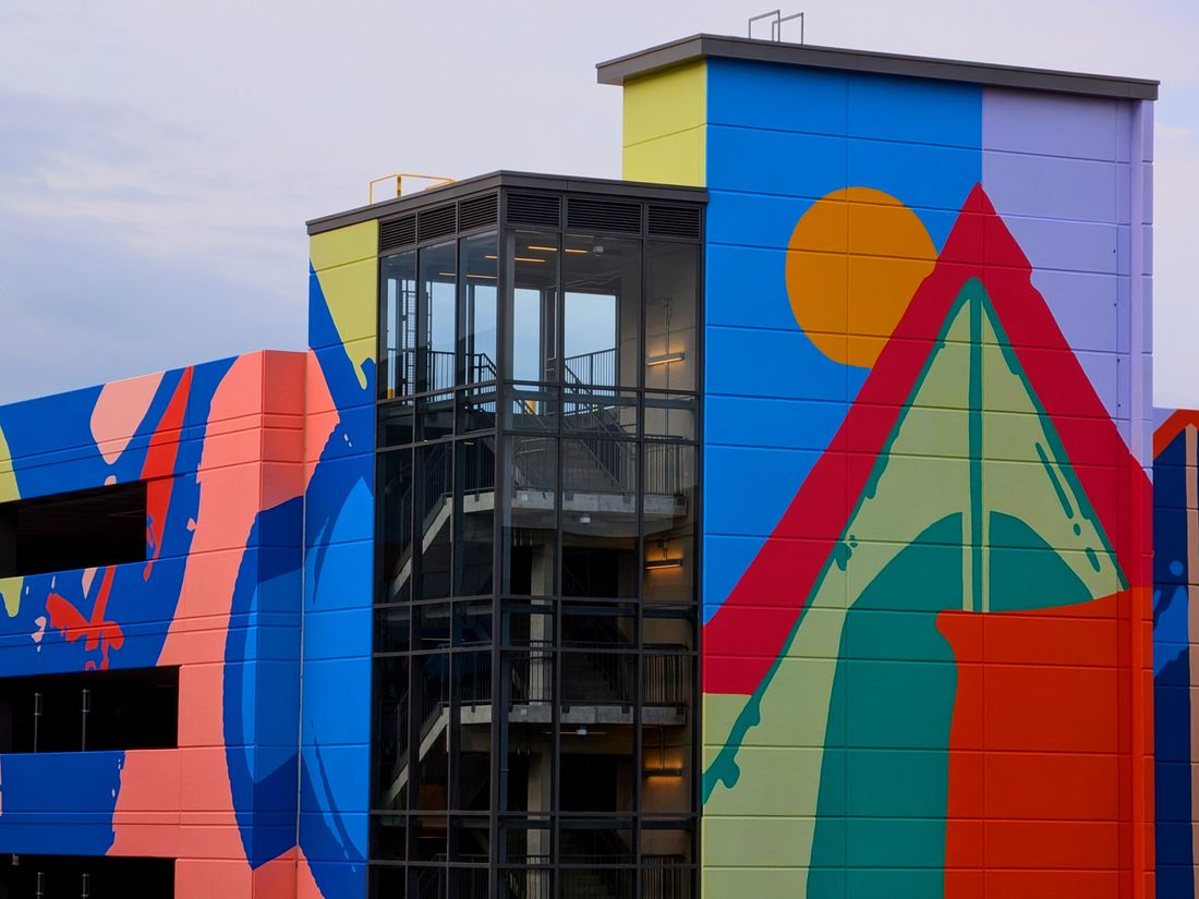 A large abstract mural in bold blue, coral, red, and yellow geometric shapes covers the exterior of a multi-story city building, with an open-air stairwell visible against a pale sky.