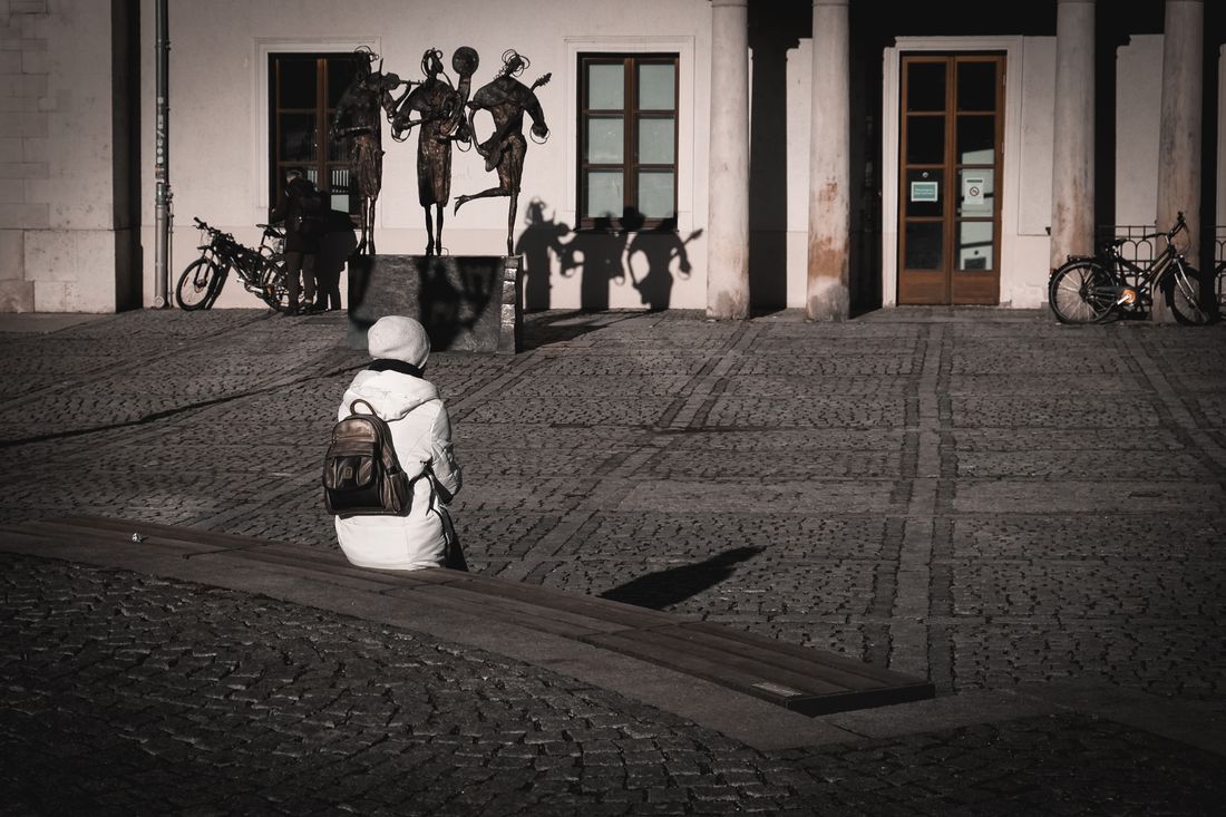 A person in a white coat sits on a bench at a distance from a group of abstract bronze figure sculptures mounted against a building wall in a quiet stone plaza, their long shadows stretching across the pavement.