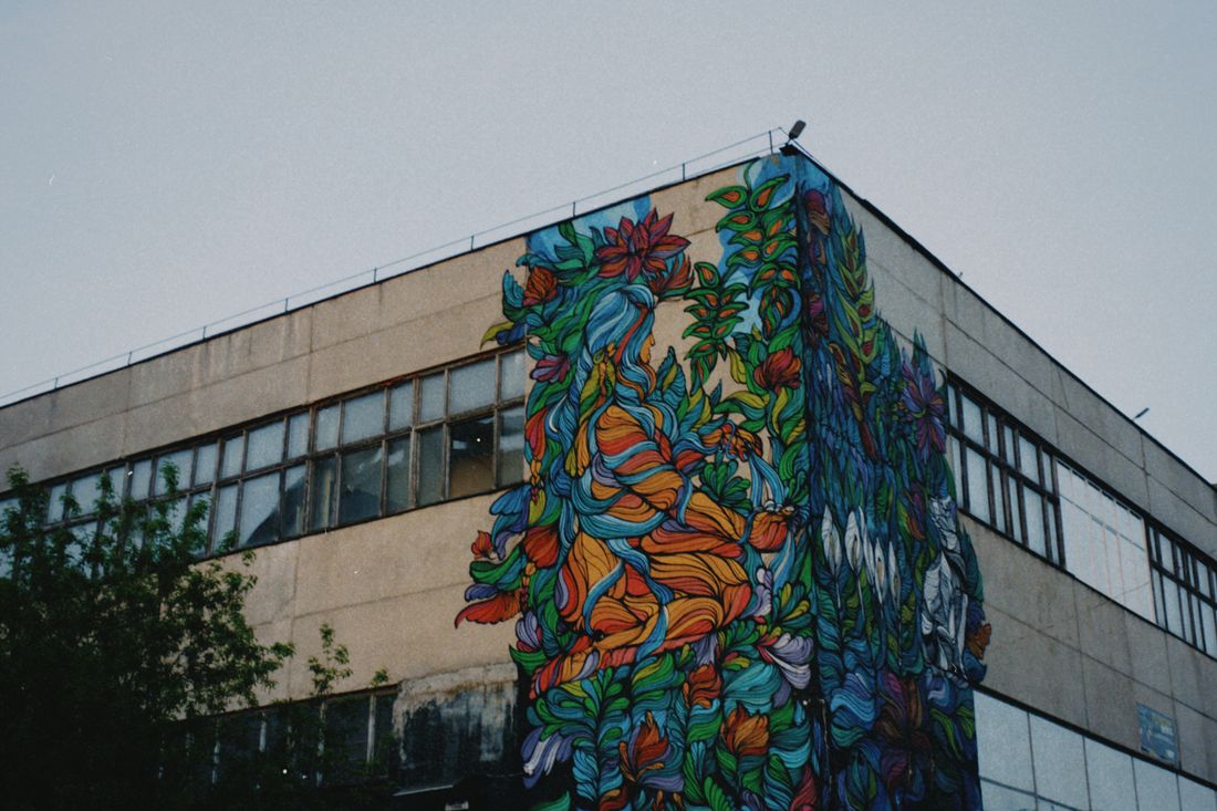 A colorful abstract floral mural wraps around the corner of a weathered concrete building exterior in soft evening light, with no people in the empty space below.