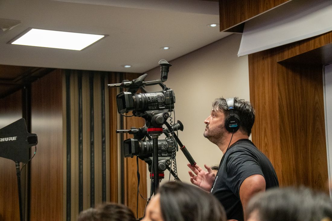 A videographer wearing headphones operates a camera on a tripod while documenting an indoor event