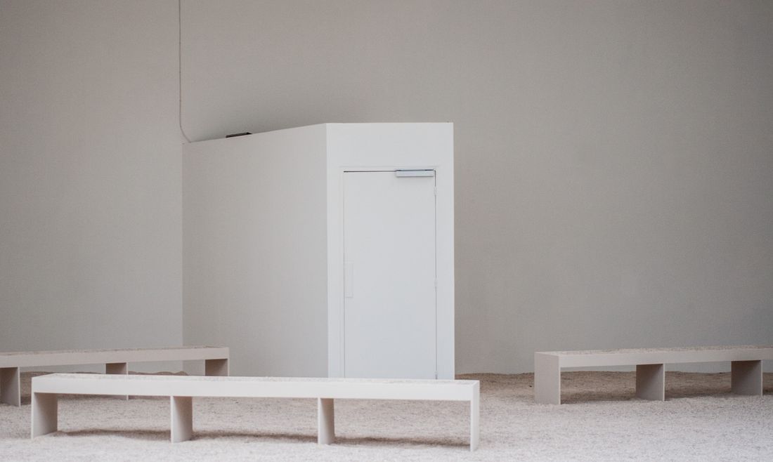 A quiet, empty gallery-style room with pale walls, a plain white structure, and long wooden benches on a pebbled floor, softly lit and free of people.