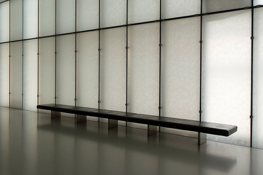 A single dark bench positioned against a tall paneled wall in an empty interior, suggesting a fixed listening or viewing spot in a quiet room.