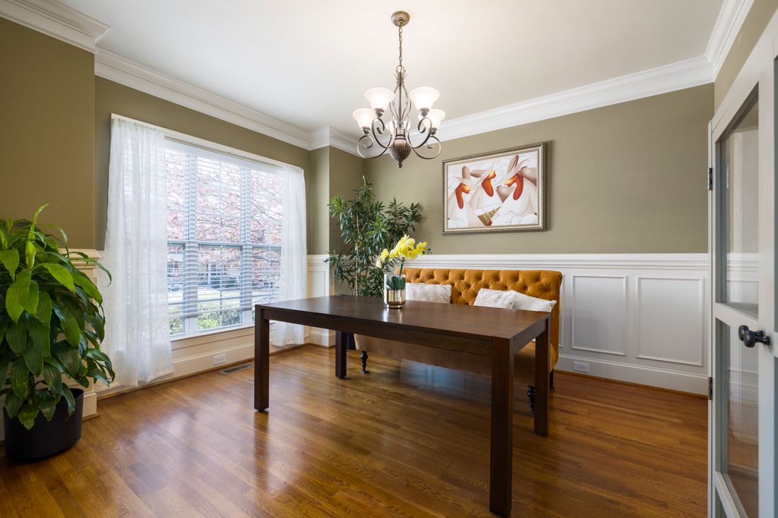 A framed abstract painting on an olive dining room wall, lit by daylight through a sheer-curtained window beside a table and tufted bench.