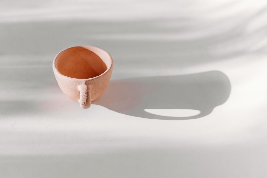A single unglazed pink earthenware cup sitting on a plain white surface in raking natural light, casting a long soft shadow.