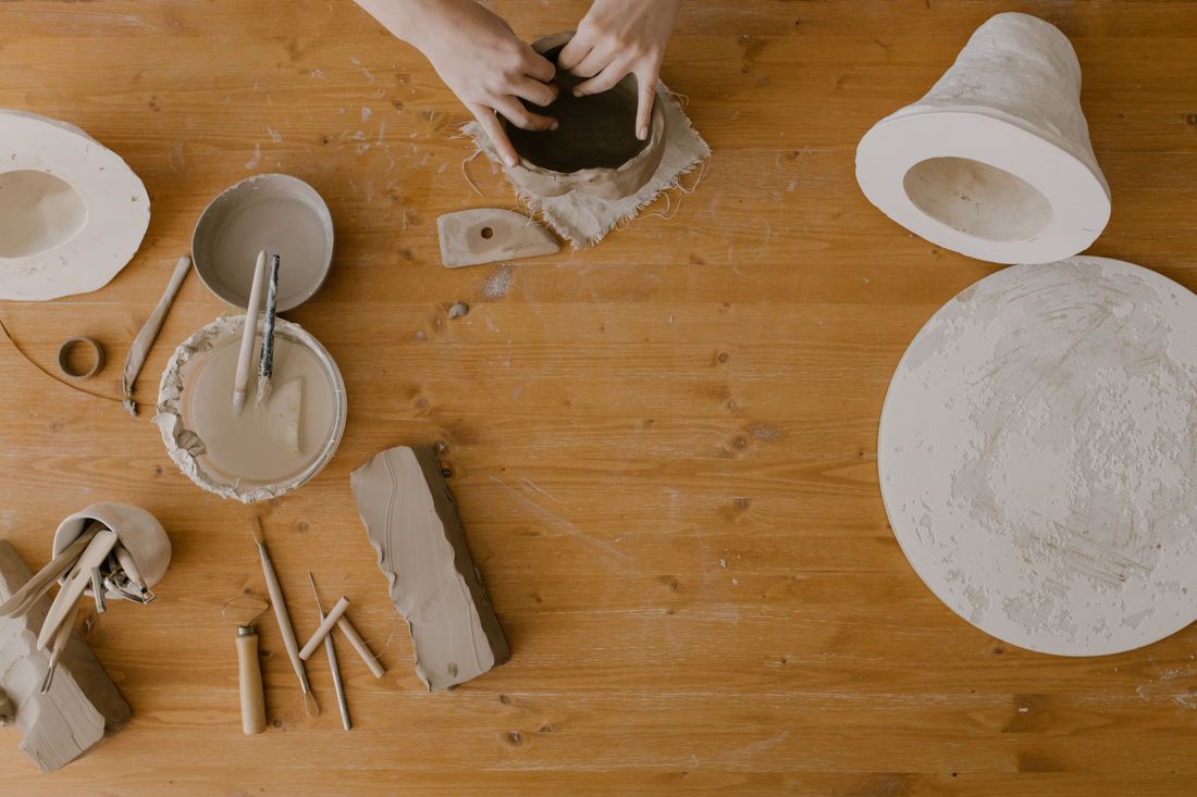 An overhead view of hands shaping clay at a wooden table surrounded by basic pottery tools and materials.