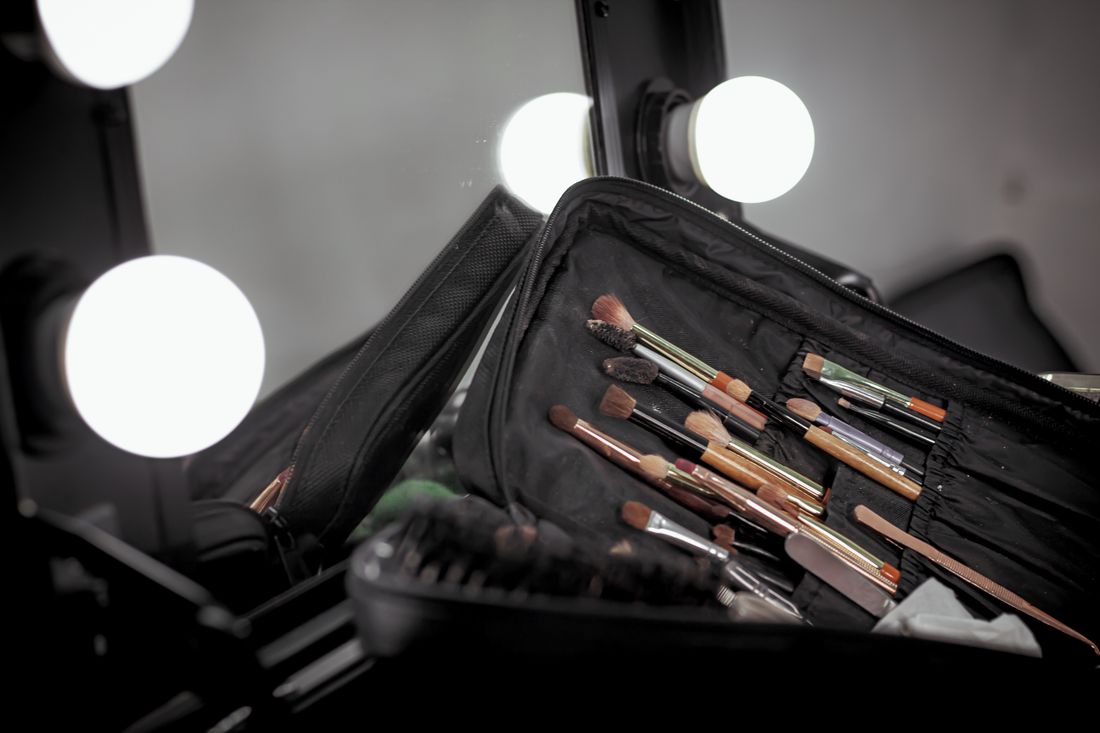 An open makeup artist's kit with brushes and tools laid out on a table, with a lit vanity mirror softly out of focus in the background.