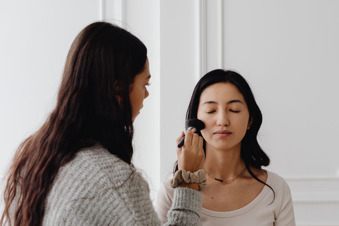 A makeup artist applies foundation with a brush to a seated woman's face in profile, against a plain wall.