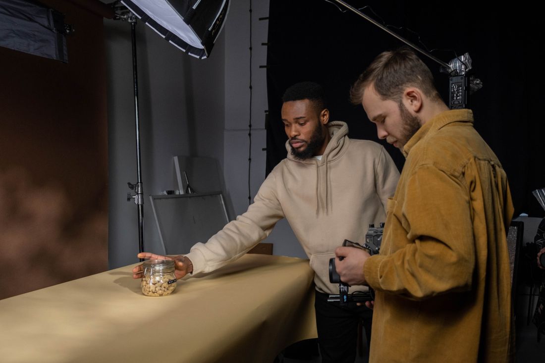 Two men in a photo studio, one placing a small jar on a table while the other holds a camera, with a softbox light on a stand nearby.