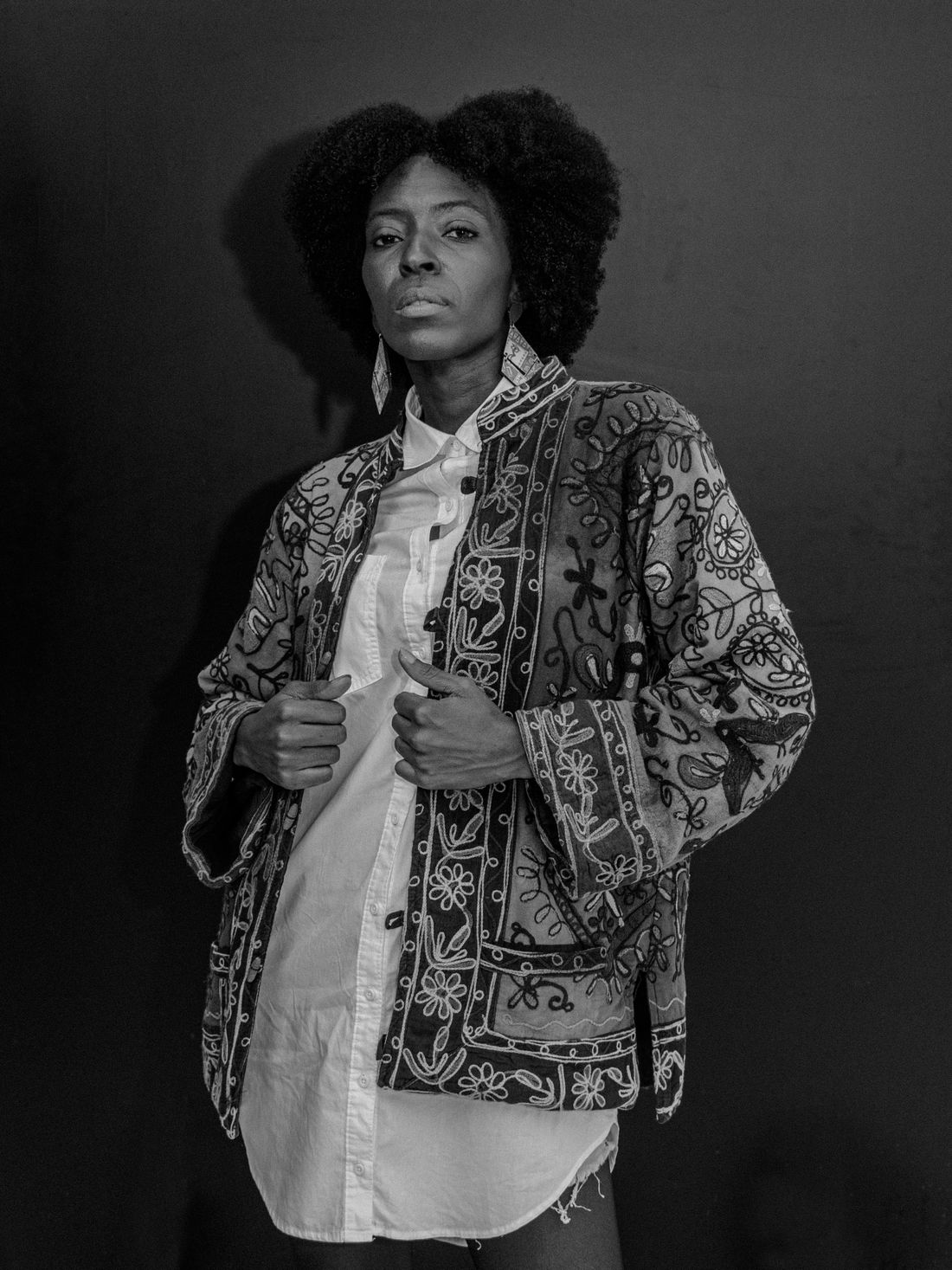 A woman in a patterned embroidered jacket standing and facing the camera against a plain dark studio backdrop