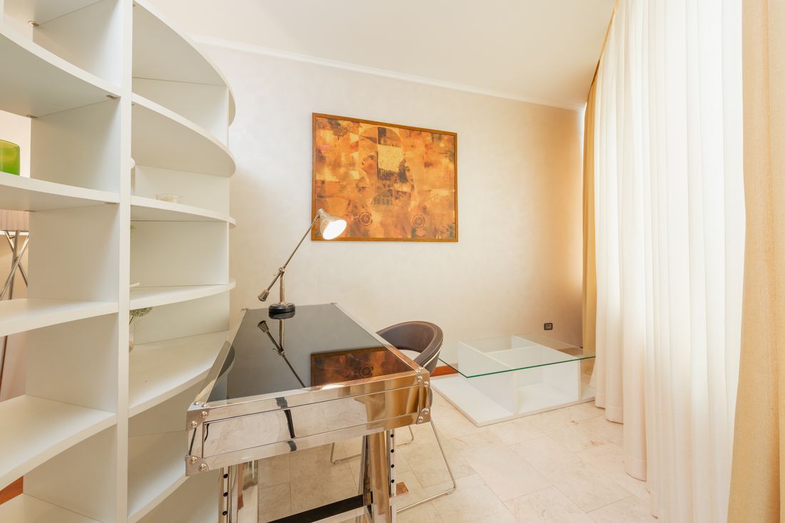 A calm, tidy workroom with open shelving, a small desk, a lamp, and a framed abstract painting, nobody present