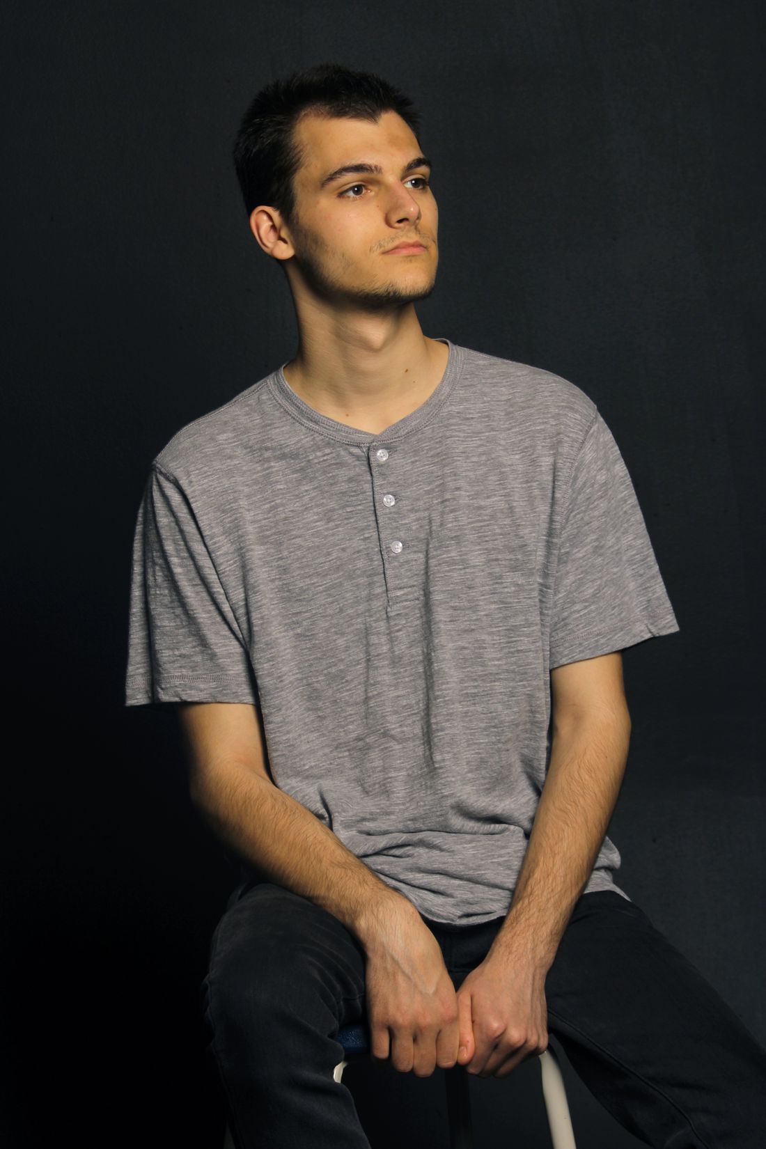 A young man seated on a stool with hands loosely clasped, wearing a gray henley, against a plain dark background