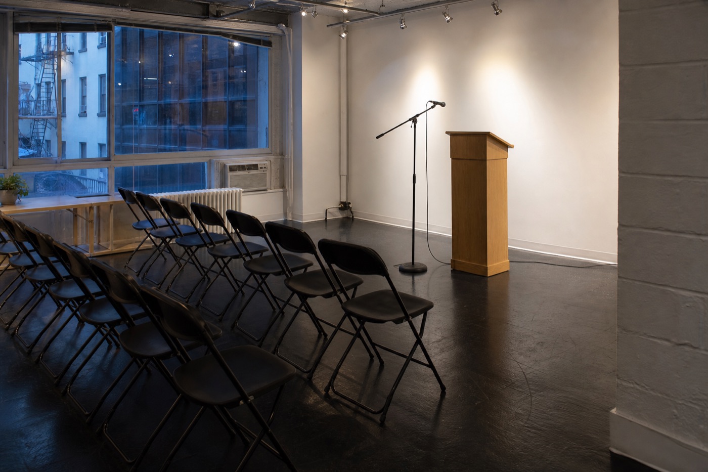 Compact event room with chairs, lectern, supported microphone, visible floor stand and cable, and a clear side aisle before guests arrive