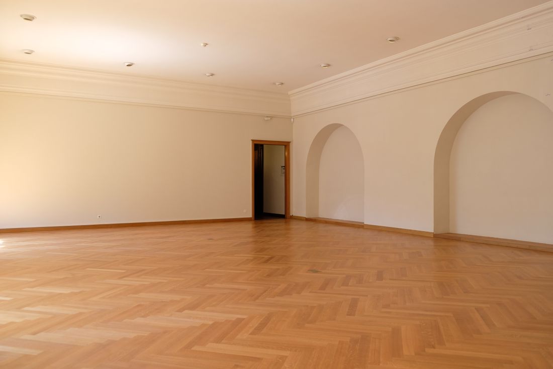 A large, empty, brightly lit room with herringbone wood flooring and blank walls, giving a long clear sightline across the open floor with two archways and a doorway in view.