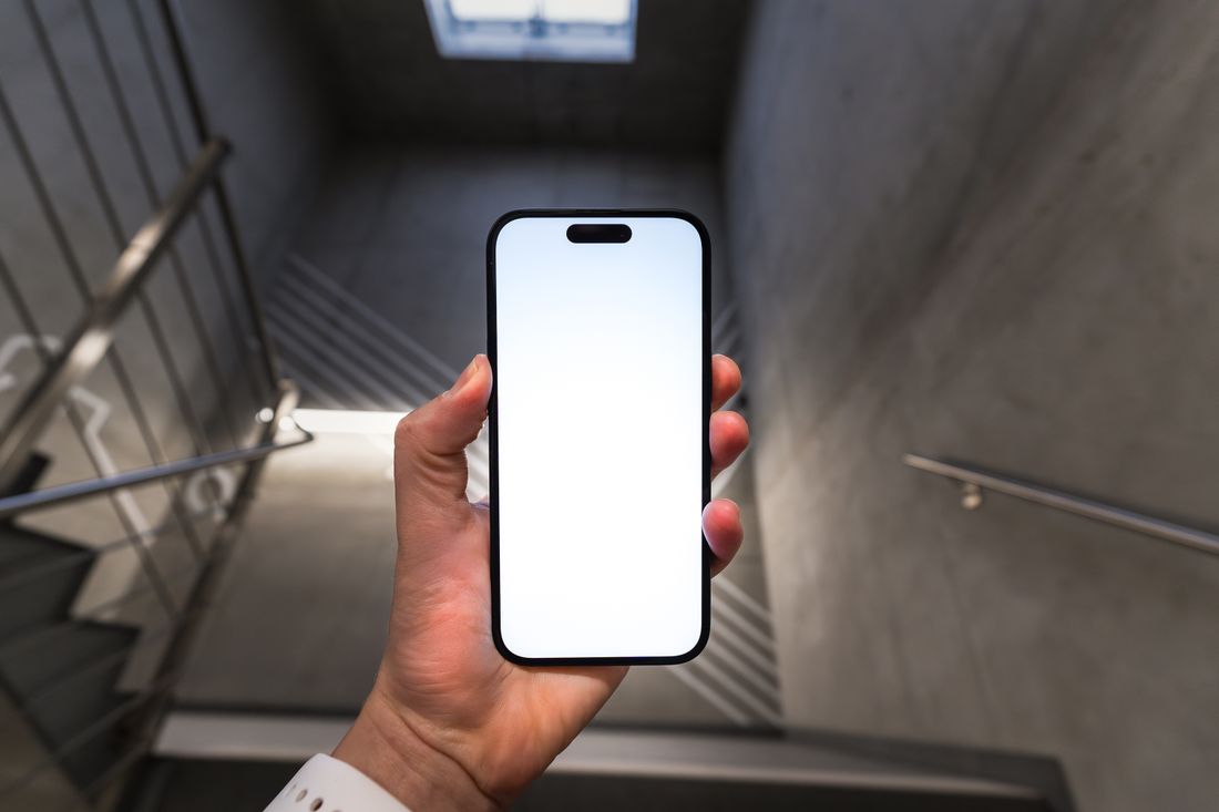A hand holding a smartphone in vertical orientation with a blank screen, cropped above the wrist with no face visible, against a blurred concrete stairwell.