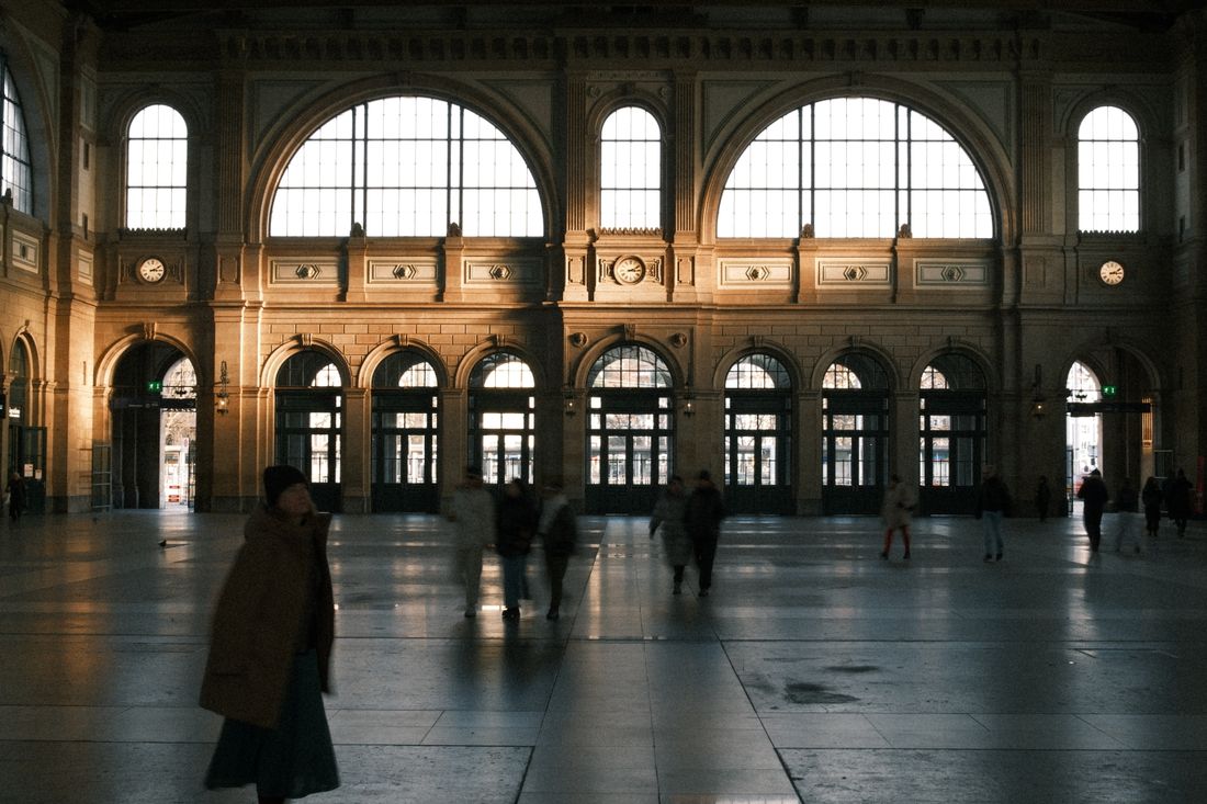 A grand arched hall with tall windows, small figures partially blurred in motion as they walk across the wide floor at a distance.