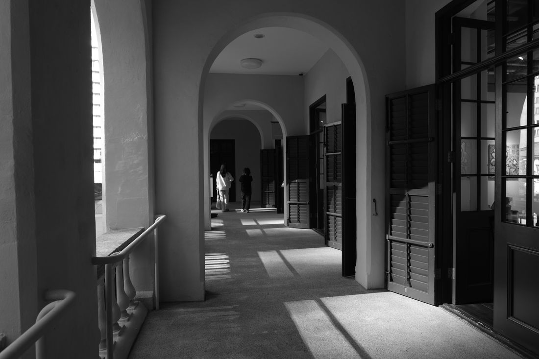 Two small figures walking away down a shadowed, arched corridor in a black-and-white photograph, doorways and window light lining the passage.