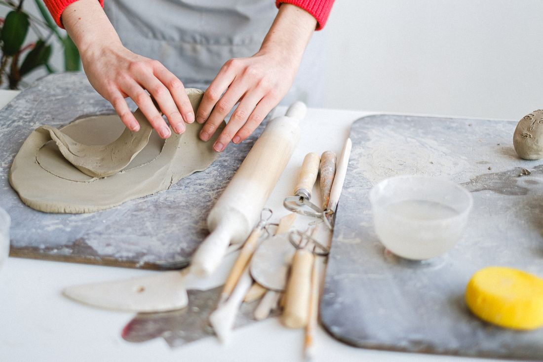 A pair of hands shaping a slab of wet clay on a workbench surrounded by pottery tools