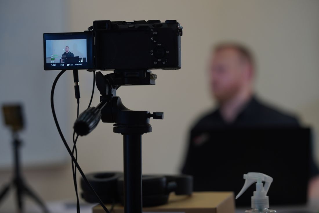 A video camera on a tripod recording a blurred speaker in the background of a quiet room
