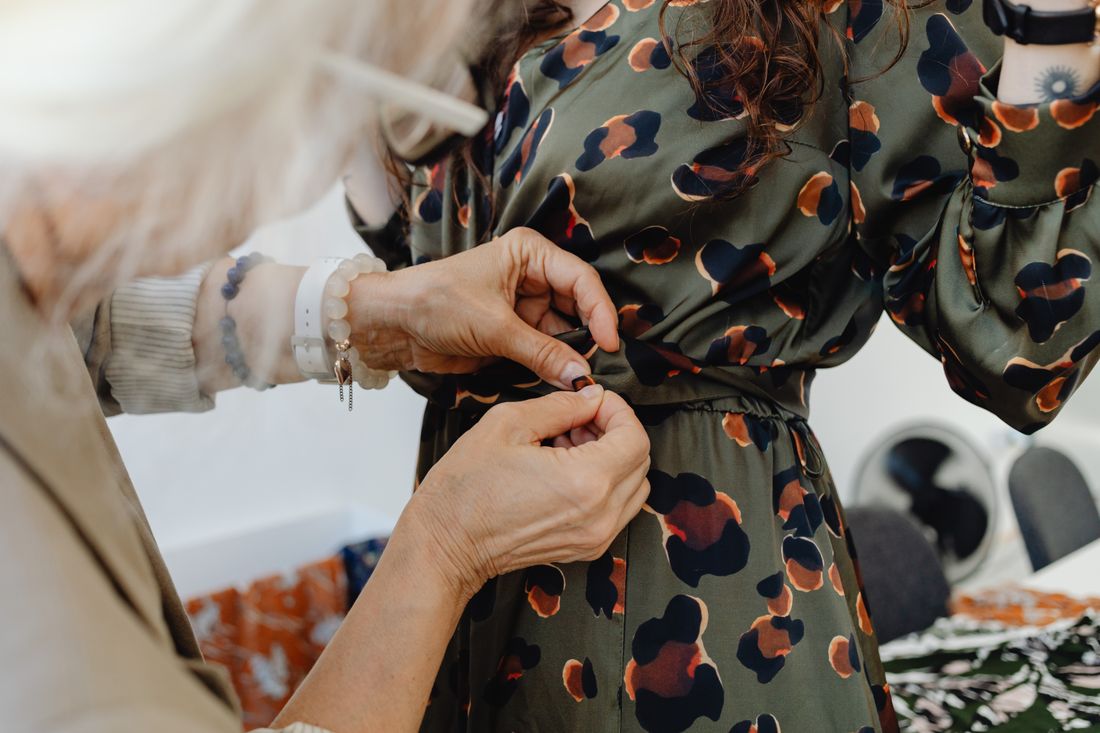 A stylist's hands adjust the back closure of a patterned dress on a subject, torso visible from waist to shoulder.