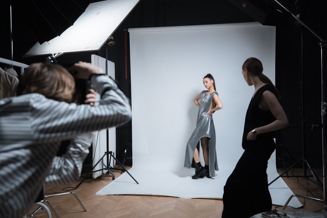 A photographer and stylist work with a subject posing against a studio backdrop, camera and lighting equipment visible.