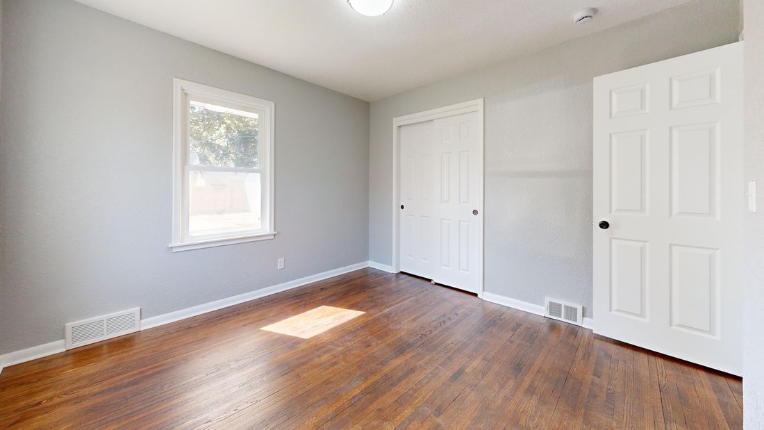 An empty room with gray walls and a hardwood floor, lit by daylight from a single window, with no furniture or fixtures in the space.