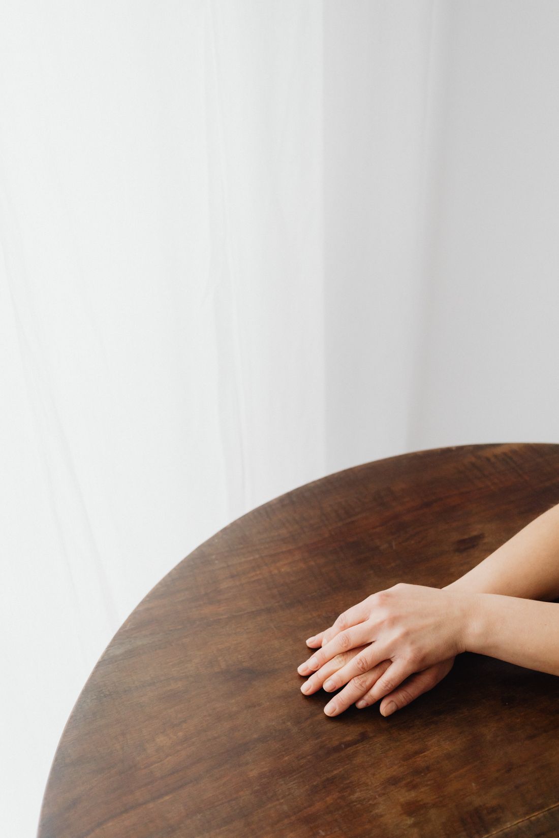 Two clasped hands resting on a round wooden table against a plain white curtain background, no face or objects in frame.