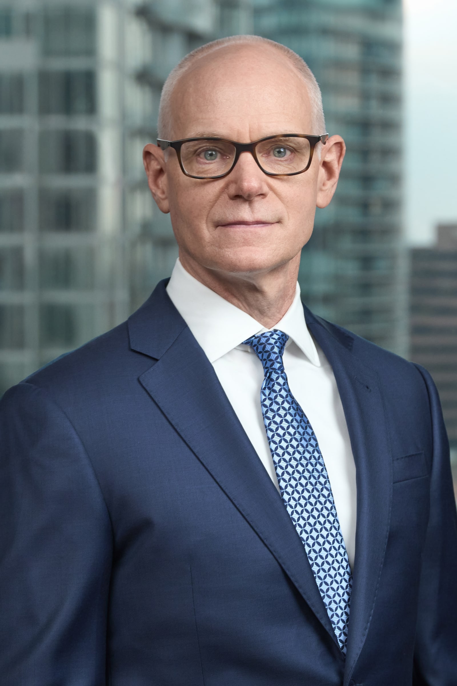 A bald man in a navy suit and glasses, photographed against a blurred city skyline, a typical corporate headshot.