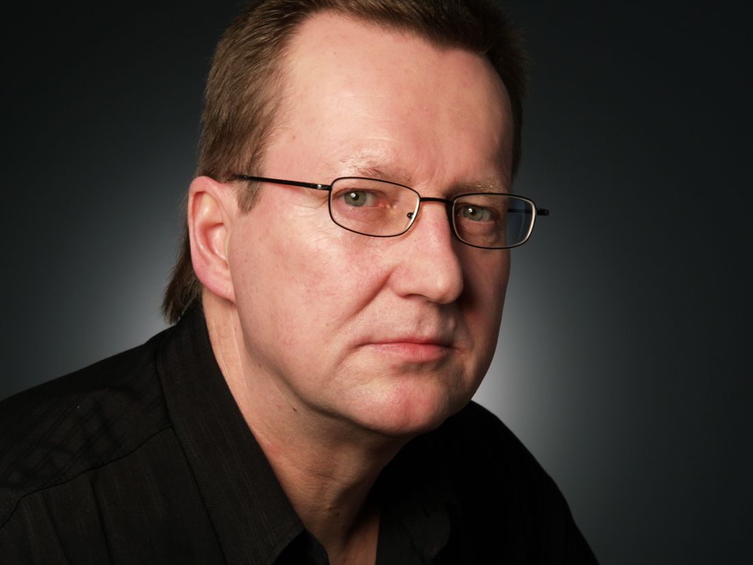 A close-up portrait of a man in eyeglasses against a plain dark studio background, a typical corporate headshot