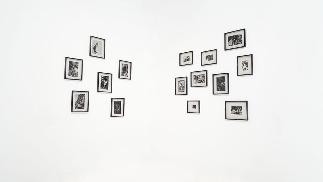 A cluster of small black-framed black-and-white photographs arranged on a plain white gallery wall, viewed from a distance with no one in the room.